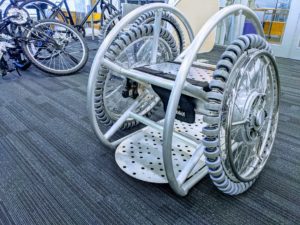 This is the image of a prototype wheelchair made in alluminium that features no backrest, but large and high metal side guards. The surface of the rear wheels is covered in small wheels positioned perpendicular to the wheel axis to allow the user to move sideways.