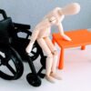 A wooden doll mimiking a transfer from a small 3d printed wheelchairs and a bench